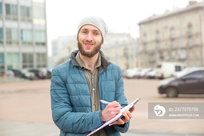 Young man makes records of research or questioning against the background of a city street. Public opinion poll, collection of statistical data, questioning.