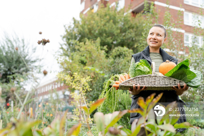 Portrait of smiling woman holding basket with fresh vegetables in urban garden