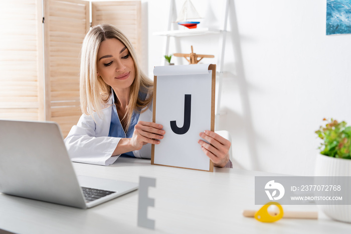 Speech therapist holding clipboard with letter during online lesson on laptop in consulting room.