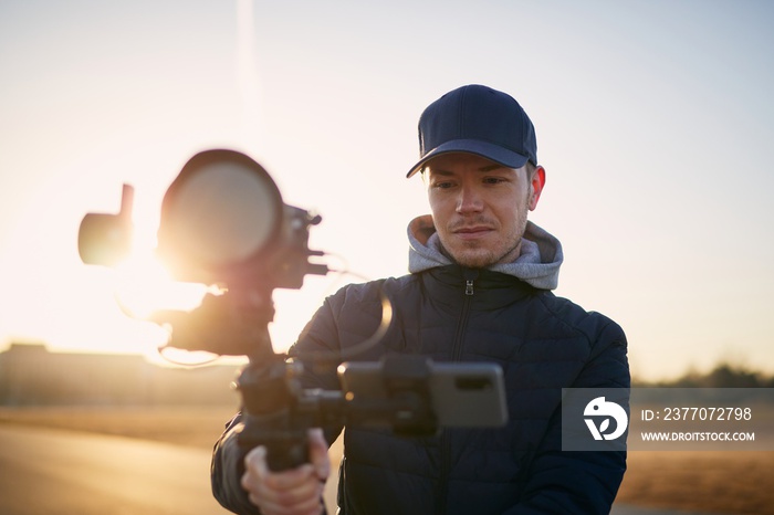 Young man filming with camera and gimbal. Portrait of videographer at beautiful sunrise.