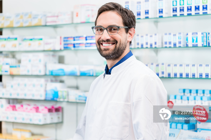 Apothecary in pharmacy standing at shelf with drugs smiling