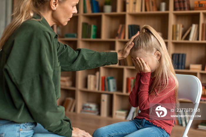 Little scared girl hiding face behind hands, ignoring caring female psychologist during personal consultation at office