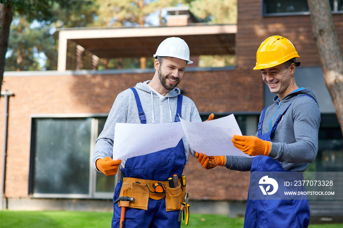 Decision making. Two positive builders smiling while standing outdoors with an open blueprint discussing the half completed cottage house