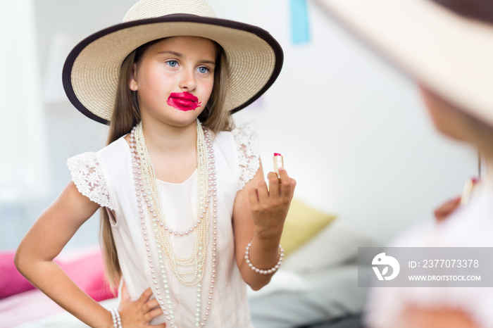 Mirror reflection shot of little girl playing dress up in mothers jewels and hat putting makeup and pink lipstick on, messing it up in smudges and looking displeased with herself