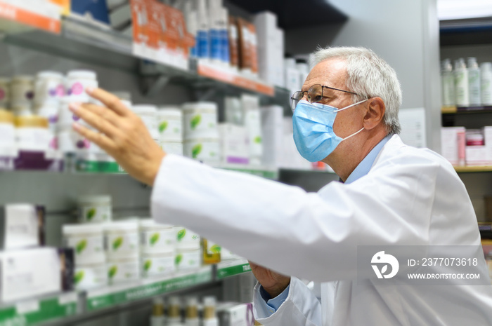 Senior pharmacist searching for a product on a shelf in his store, wearing a mask due to coronavirus