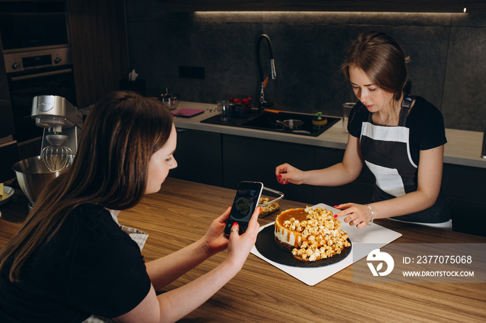 Young woman taking pictures of a cake in the kitchen. Female baker capturing photos of pastry items with her mobile phone.