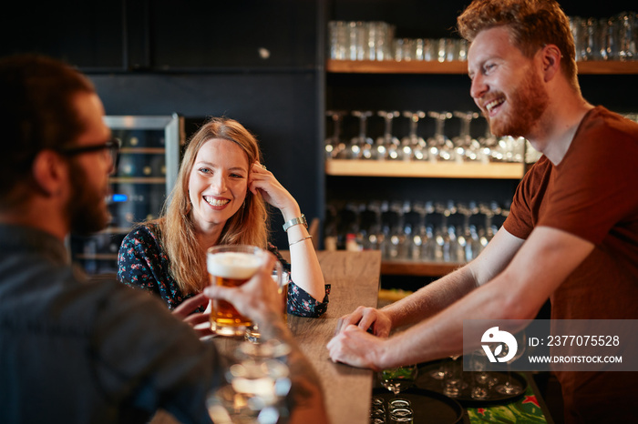 Cheerful friends leaning on bar counter, drinking beer and chatting with bartender. Night out.