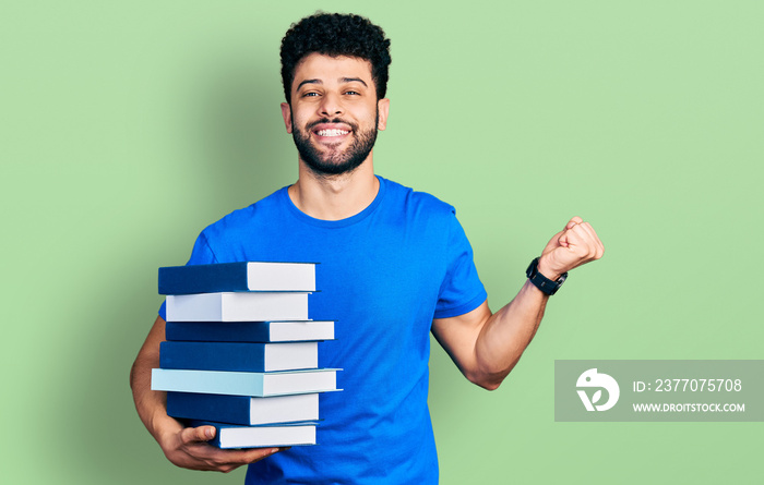 Young arab man with beard holding a pile of books screaming proud, celebrating victory and success very excited with raised arm