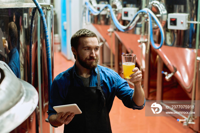 Happy young brewer with digital tablet looking at glass of beer in his hand after preparation while standing in aisle against huge steel tanks