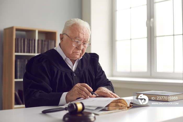 Wise senior lawyer in glasses working with case documents in office. Serious mature judge in gown uniform reading book while sitting at table in court of law. Concept of judicial system and justice