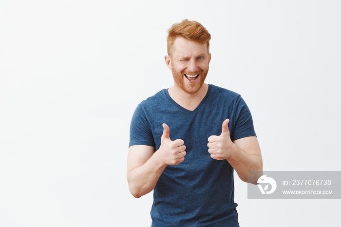 Yeah boy nice choice. Portrait of good-looking male friend with ginger hair showing thumbs up and winking with hint, liking and supporting great decision, cheering over gray background