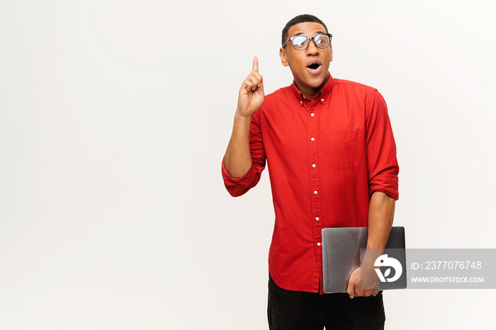 Young African-American short-cutted student in glasses holds laptop and finger point up isolated on white background, guy in red shirt excited with breakthrough. Tech geek come with task solution