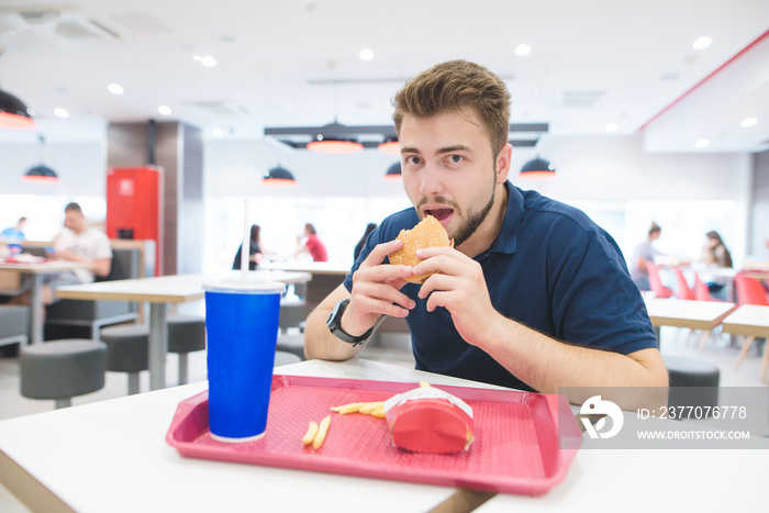 Man with a beard sits at a table with a tray in a fast-food restaurant with a burger in his hands and looks at the camera. Student is eating fast food at the fast food restaurant.