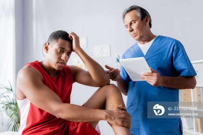 chiropractor holding digital tablet near upset african american man in sportswear in clinic