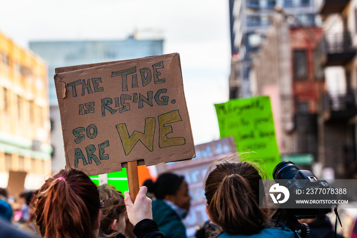 Activists unite for climate change. A closeup view of a cardboard sign reading the tide is rising, so are we, above a gathering of environmental demonstrators on a city street.