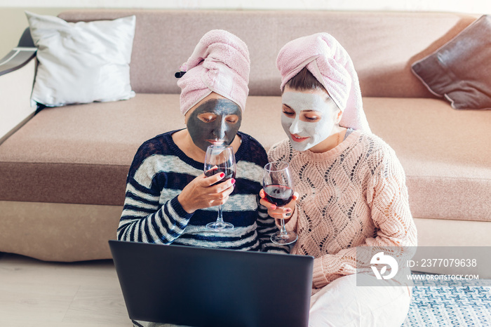 Mother and her adult daughter drinking wine and using laptop with facial masks applied. Women chilling at home