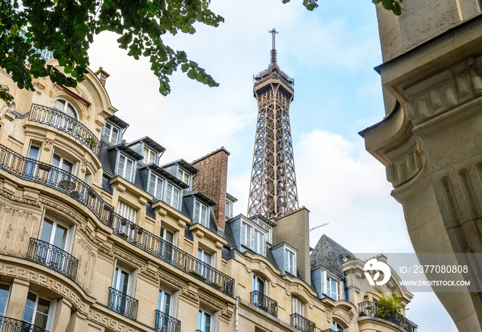 The top of the Eiffel Tower seen from down the street with foliage and typical residential buildings in the foreground.