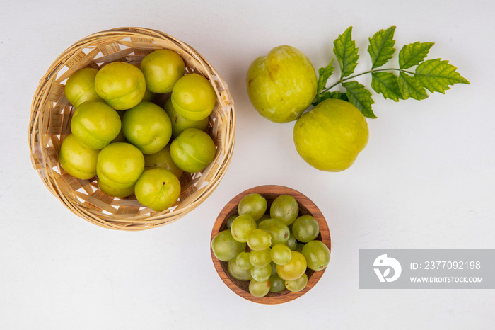 top view of fruits as plums in basket and green pluots with bowl of white grape berries on white background