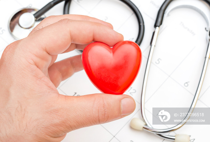 Man’s hand holding red heart over calendar and stethoscope.