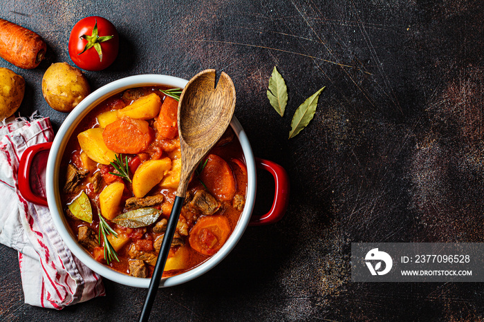 Slow cooked beef stew with potatoes and carrots in tomato sauce in pot, dark background.