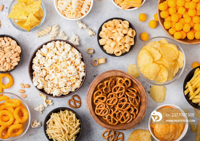 All classic potato snacks with peanuts, popcorn and onion rings and salted pretzels in bowl plates on light background. Twirls with sticks and potato chips and crisps with nachos and cheese balls.