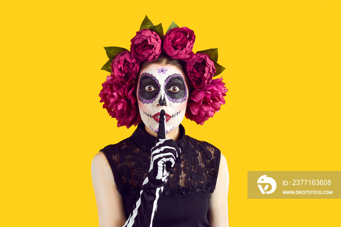 Woman with sugar skull makeup and wreath of beautiful roses on head standing on yellow studio backgr