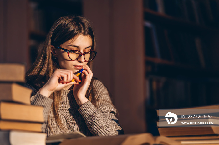 Young student in glasses preparing for the exam. Girl in the evening sits at a table in the library 