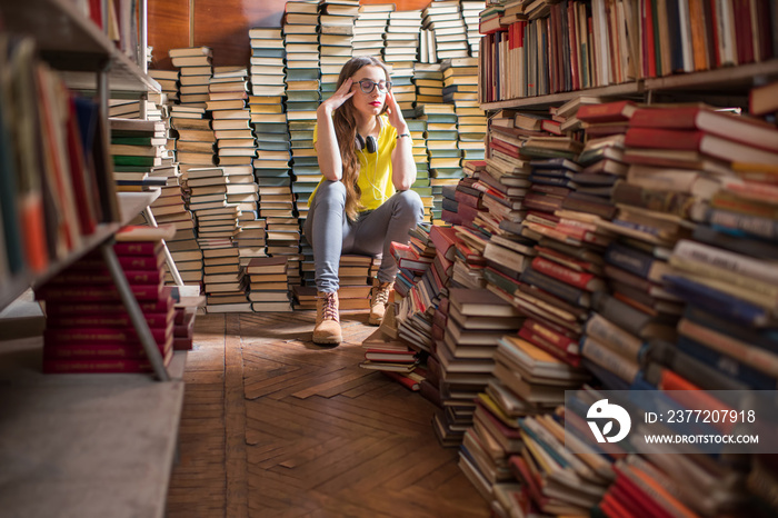 年轻疲惫的女学生坐在旧图书馆的一堆书上