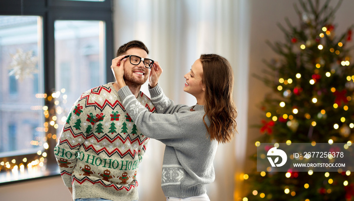 christmas, people and holidays concept - happy couple in ugly sweaters over home background