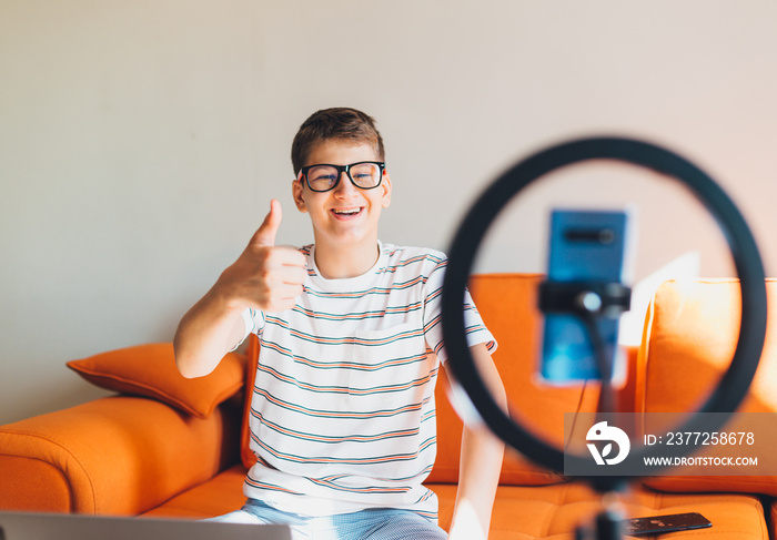 Young teenager blogger in glasses recording video at home. Cheerful cute boy pointing with hands int