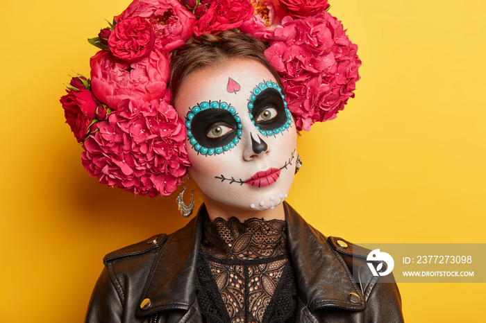 Close up shot of mysterious woman with skull makeup wears flower wreath and black leather jacket pre