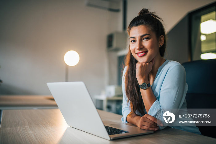 Portrait of a cute millennial girl sitting at desk with laptop, smiling at camera.