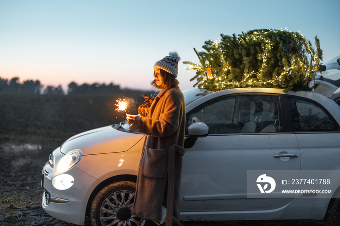 Happy caucasian woman firing sparklers near car with Christmas tree on nature at dusk. Concept of ce