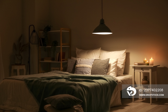 Interior of bedroom with green blankets on bed, burning candles and glowing lamp at night