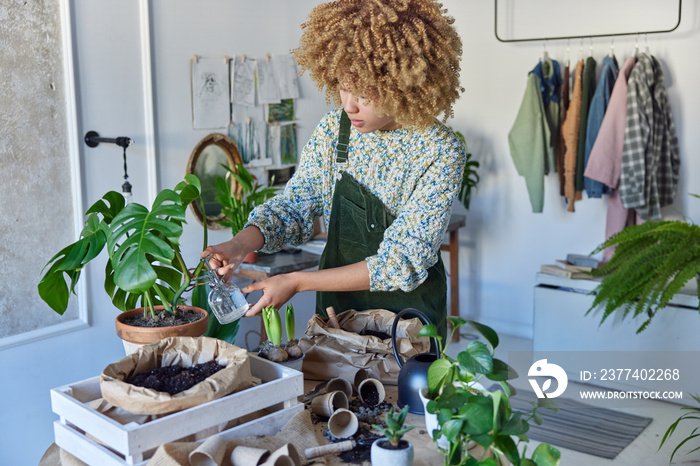 Indoor shot of curly haired woman transplants houseplants relocates flowers and sprays with water we