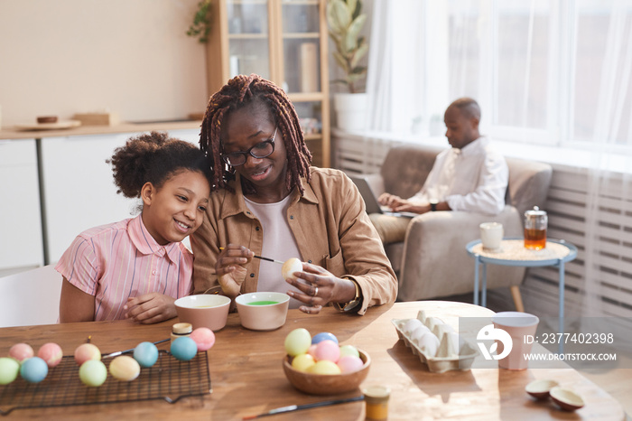 Portrait of loving African-American mother and daughter painting Easter eggs together while sitting 