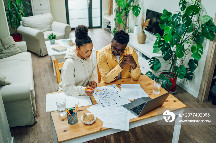 Multiracial couple using laptop and blueprints of their new home