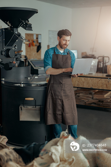 Male worker on small coffee factory working laptop on background of coffee roasting machine