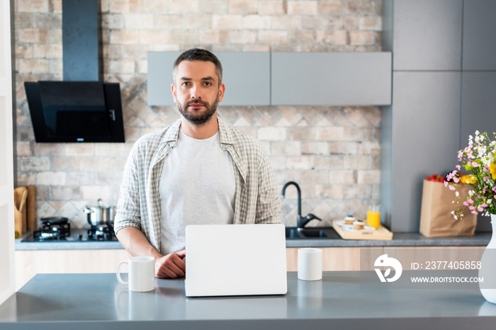 portrait of bearded man standing at counter with laptop and cups of coffee and looking at camera in 