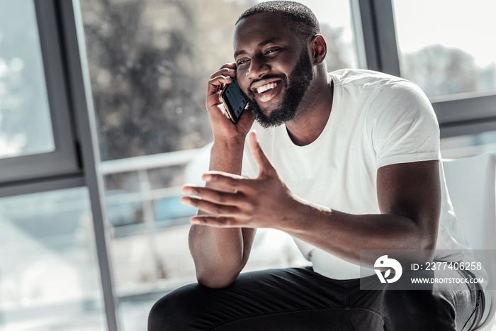 Phone conversation. Happy cheerful young man smiling and talking on the phone while sitting in the o