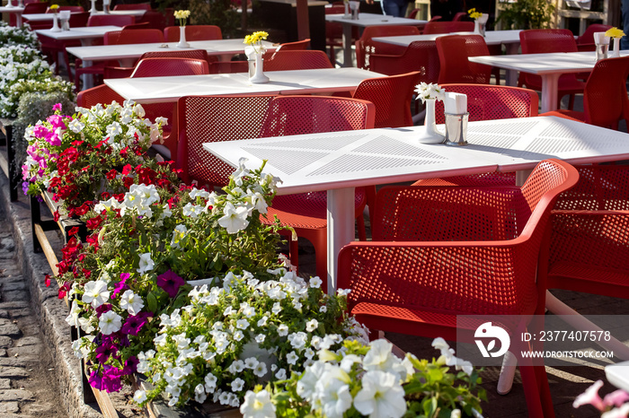 white perforated tables with red chairs of an outdoor street cafe with vases and blooming petunias o