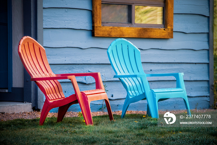 Close-up of red and turquoise arondiack chairs sitting outside blue wooden cabin door on grass.