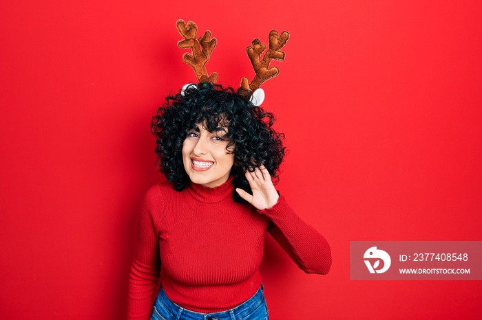 Young middle east woman wearing cute christmas reindeer horns smiling with hand over ear listening a