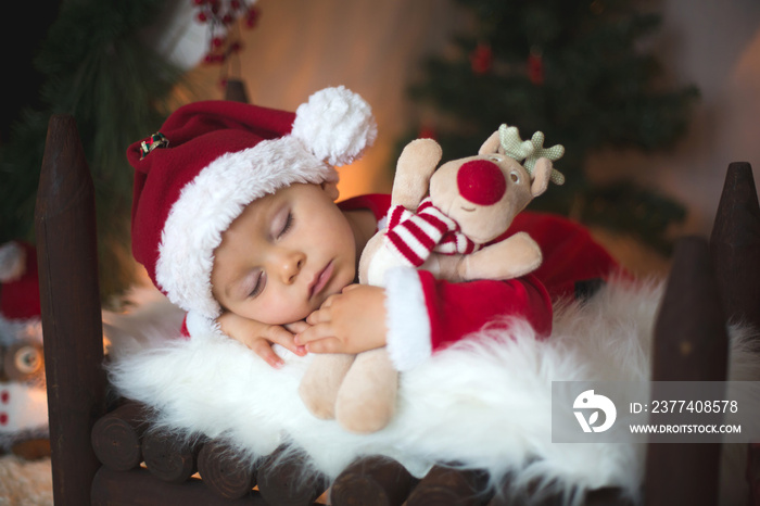 Adorable little toddler baby boy dressed in canta claus costume, sleeping in baby bed in front of cr