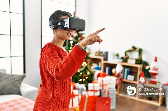 Young hispanic woman playing video game using vr glasses standing by christmas tree at home