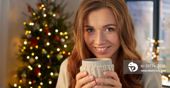 holidays and people concept - happy young woman with cup of coffee or tea at home over christmas tre