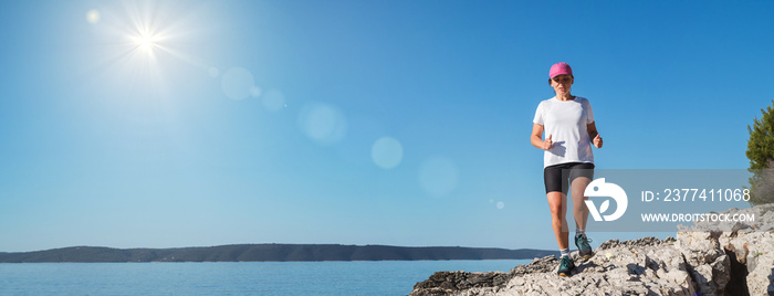 Middle-aged female dressed running sporty clothes and pink cap enjoying morning jogging along the rocky calm sea coast. Sporty people trail running activities on vacation time concept image.