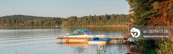 Summer on lake cottage. Small yacht boat by wooden dock pier on lake at sunset. Canadian Ontario Muskoka countryside travel destination. Summer water sport outdoor activity. Web banner header.
