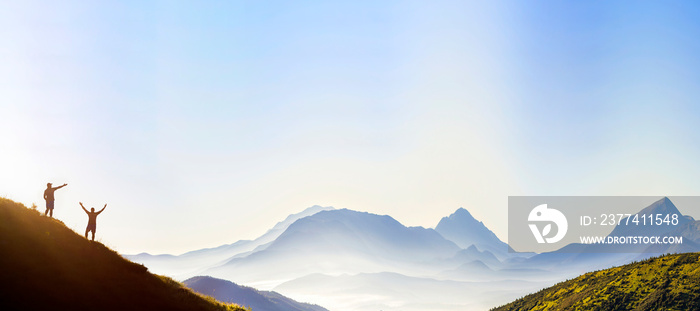 Small dark silhouettes of tourist travelers on steep mountain slope at sunrise on copy space background of valley covered with white puffy clouds and bright clear sky.