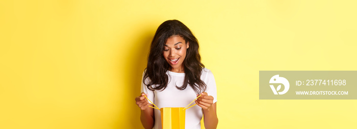 Image of happy and surprised african-american girl, receiving a gift, looking inside shopping bad with amazement, standing over yellow background